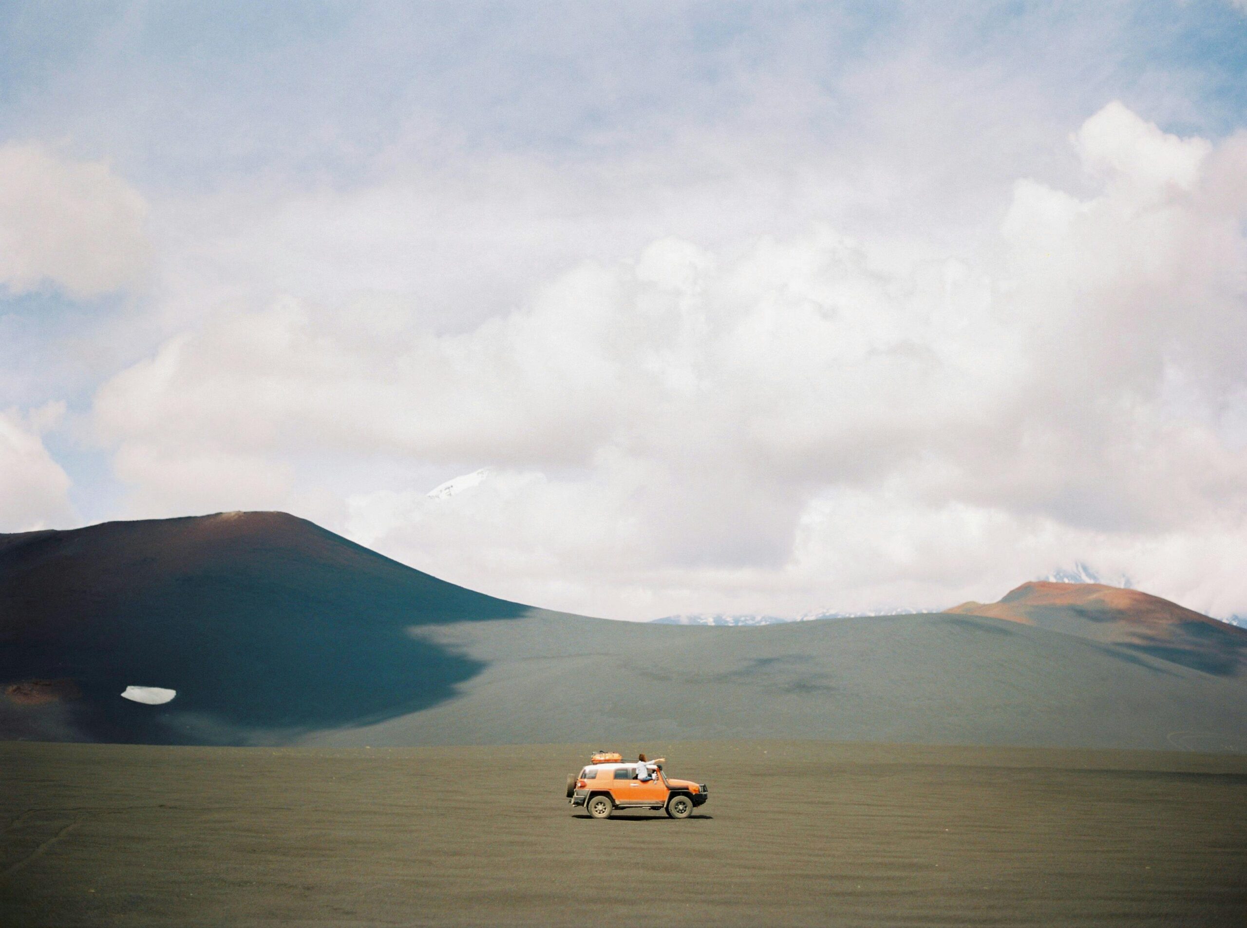 Orange off-road SUV driving through a vast volcanic plain with distant snow-capped mountains and cloudy skies.


